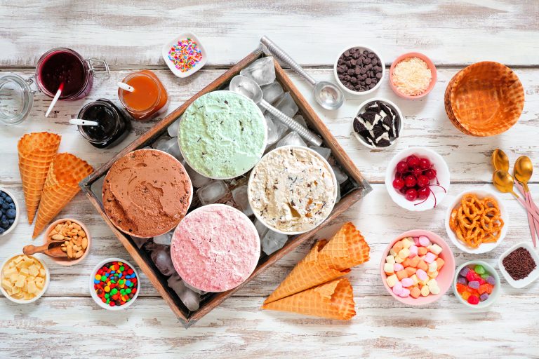 A variety of ice cream flavors in bowls, surrounded by toppings, cones, and sauces on a wooden table.
